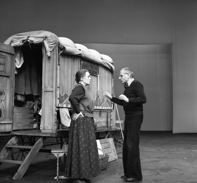 An actress in a long skirt and a man in a dark sweater talking beside a covered canvas wagon on a bare stage