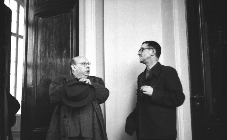 Hanns Eisler, holding a hat, talking with Bertolt Brecht beside a tall doorway