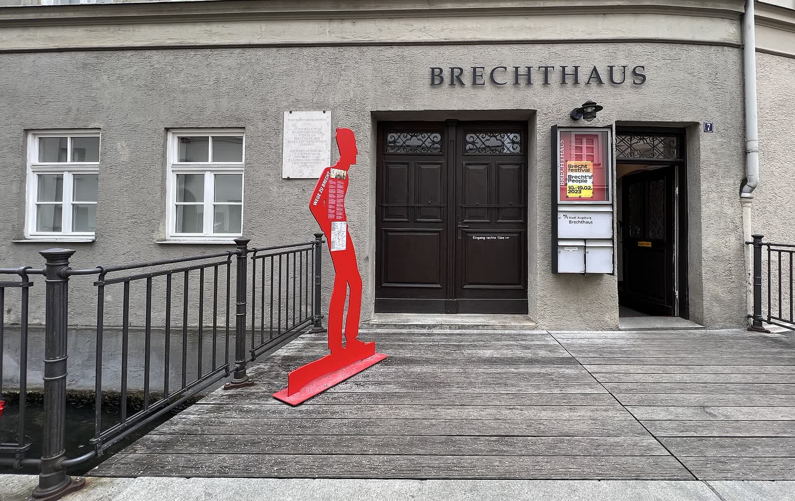 Grey facade of the Brechthaus in Augsburg with a red cut-out figure of Brecht on the footbridge to its door