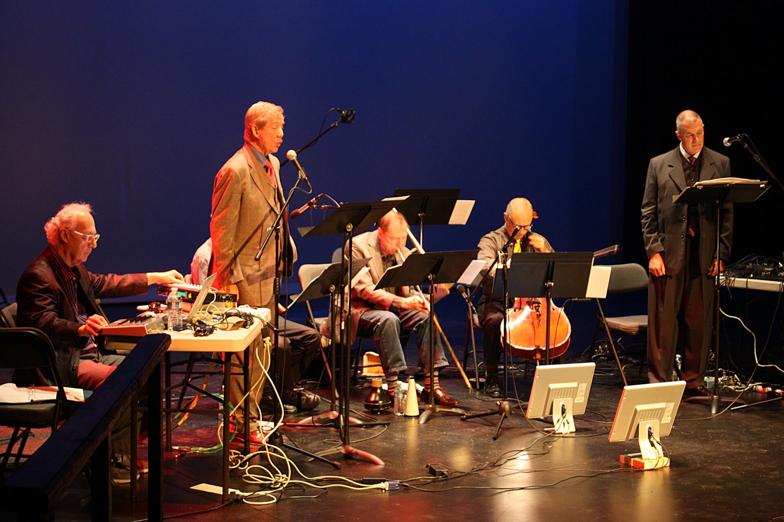 Five musicians with laptops, electronics, a cello and music stands performing on a dark stage under blue light