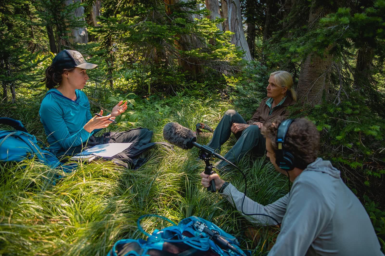 Three people sitting in a forest while one holds a microphone and wears headphones to record an interview