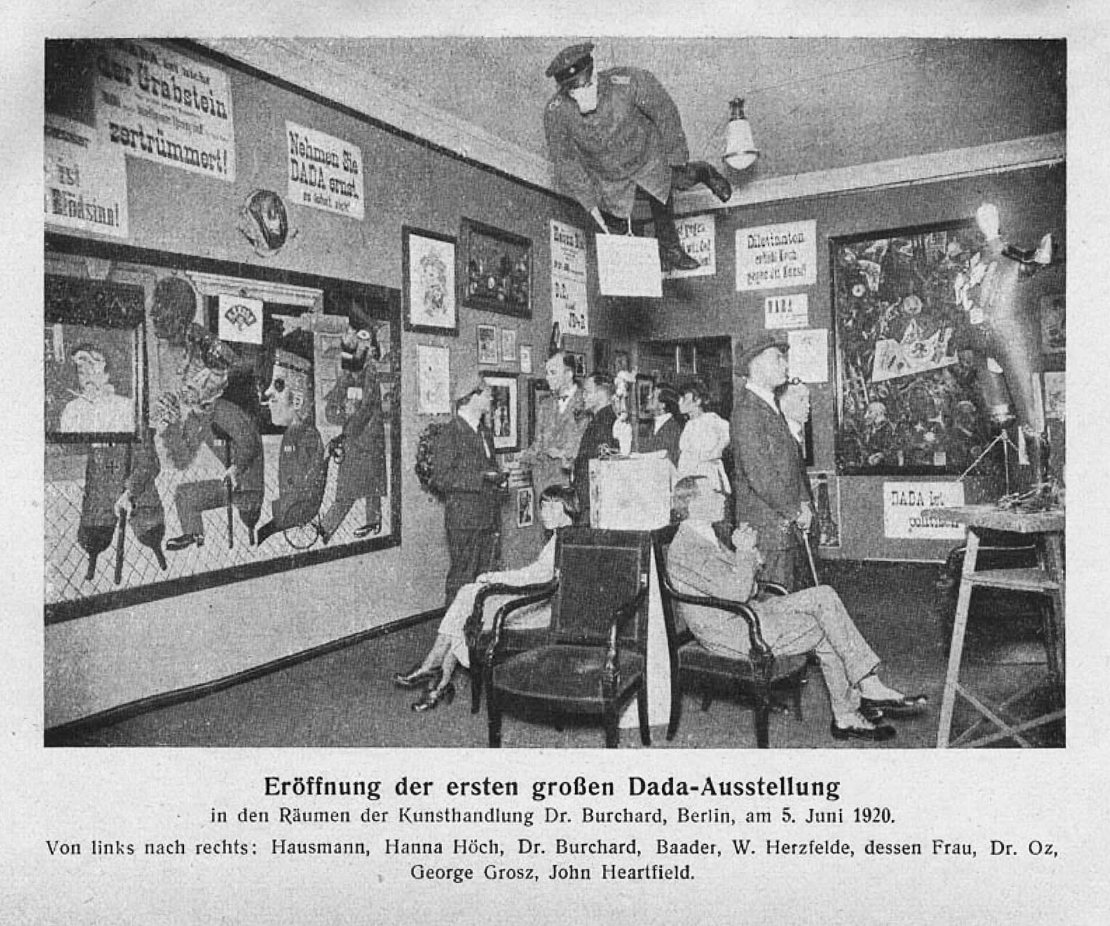 A crowded gallery room hung with posters and collages, a dummy in uniform hanging from the ceiling and visitors seated below