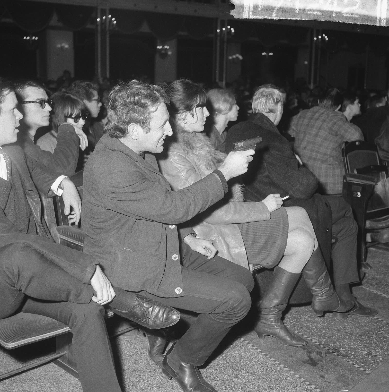 Black and white photo of a seated audience, one young man pointing toward the stage with others laughing beside him