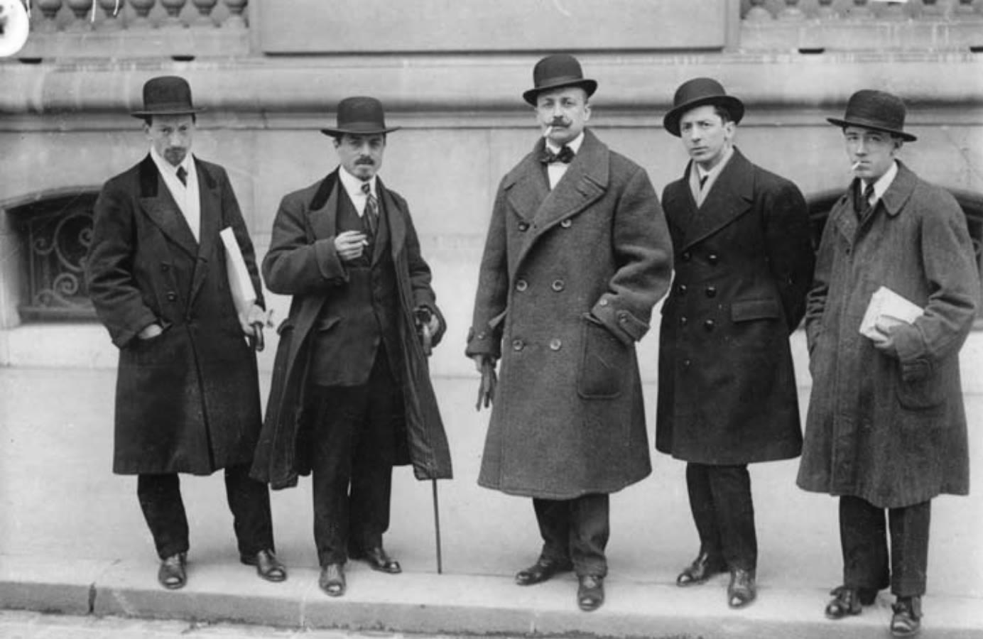 Five men in overcoats and bowler hats standing in a row on a Paris pavement