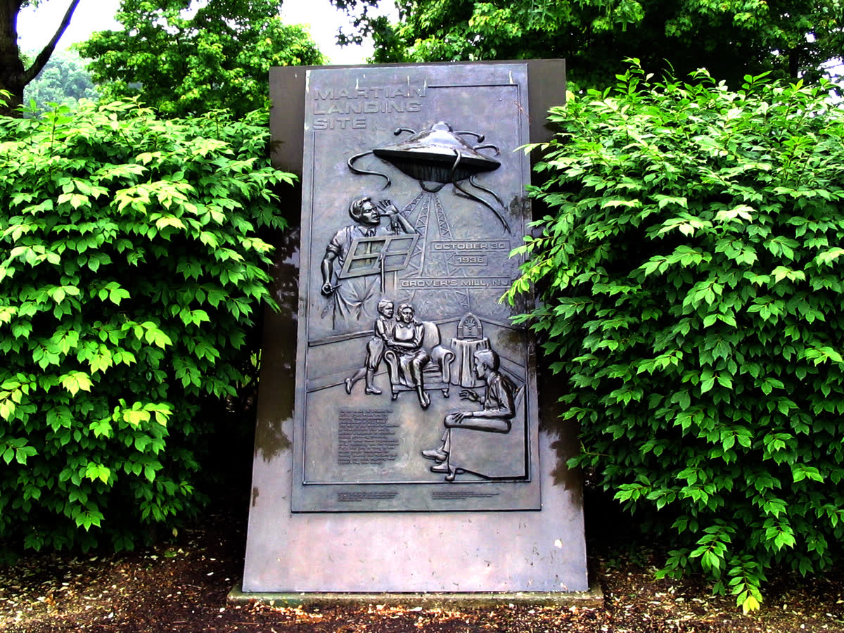 Bronze relief monument showing a Martian machine, a radio listener and Welles at a microphone, framed by green bushes
