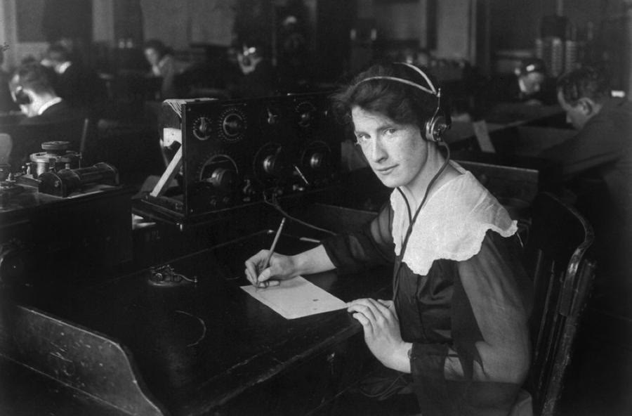 Black and white photo of a woman wearing headphones at a radio receiving desk, writing a message