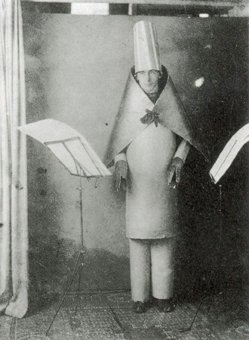 Hugo Ball standing in a stiff cardboard costume with a tall hat and a cape, between music stands