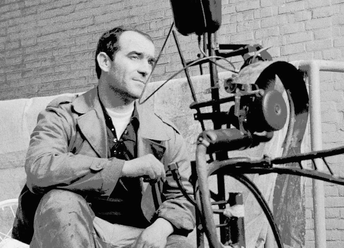 Black and white photo of Jean Tinguely sitting beside one of his wheeled metal machines