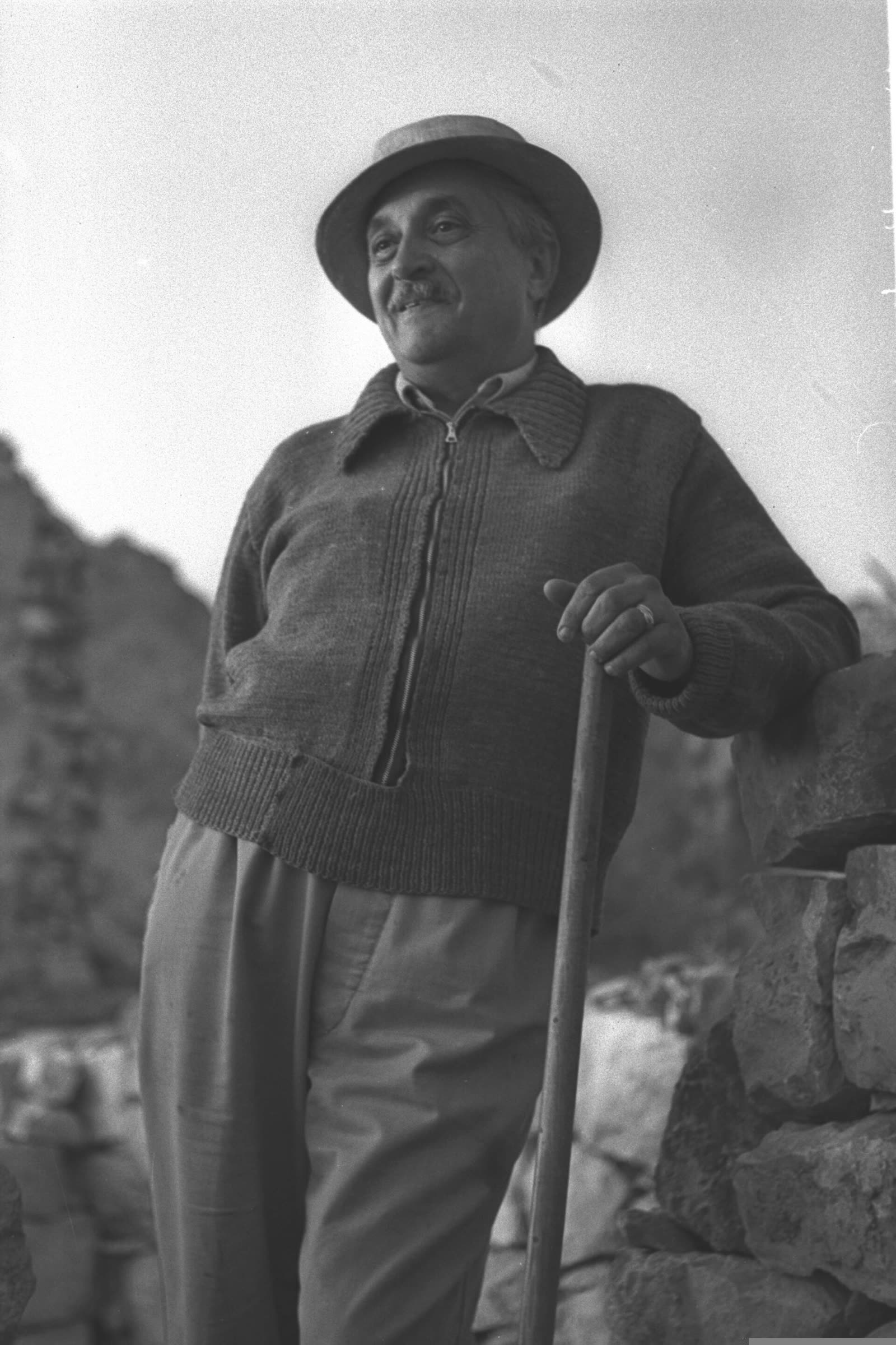 Black and white photo of an older Marcel Janco in a hat and sweater, leaning on a stick outdoors