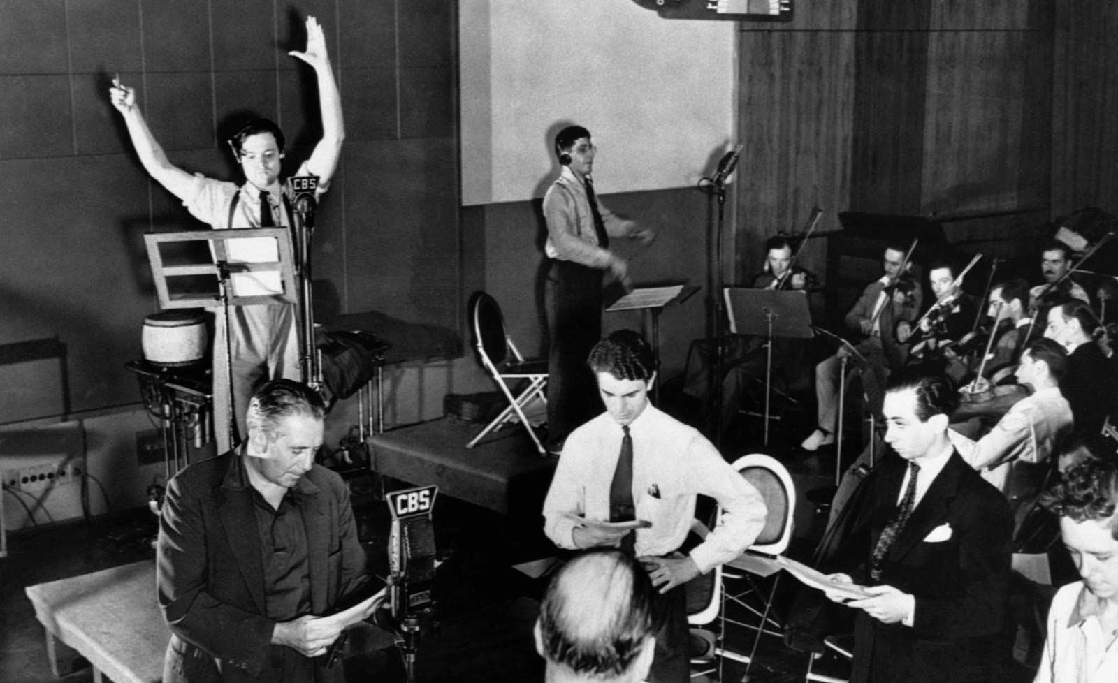 Black and white photo of Orson Welles with arms raised directing actors and an orchestra in a radio studio