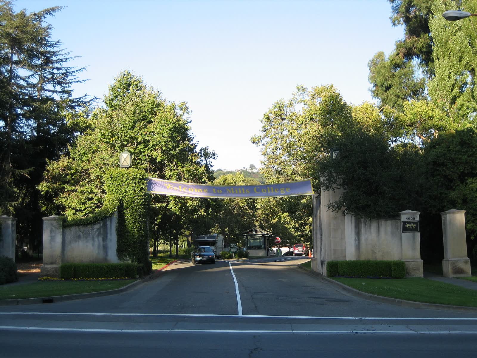 Road leading through the stone gateposts of the Mills College entrance, with a banner overhead and trees on both sides