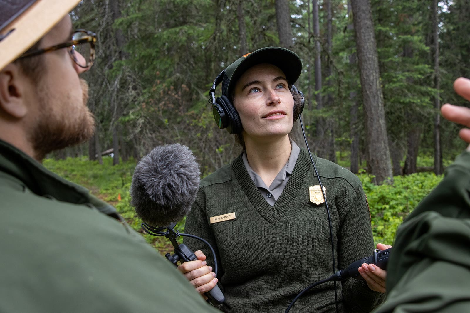 Podcast host holding a microphone and recording equipment in a forest at Glacier National Park