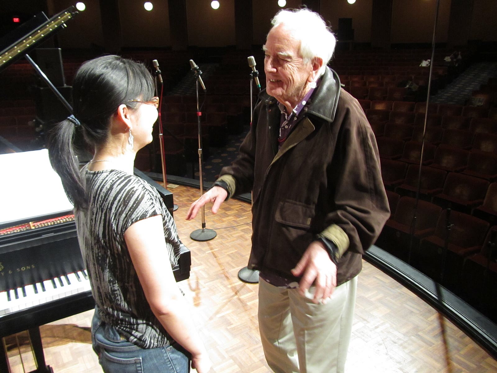 White-haired composer Robert Ashley in a brown jacket talking with a performer beside a grand piano on a stage
