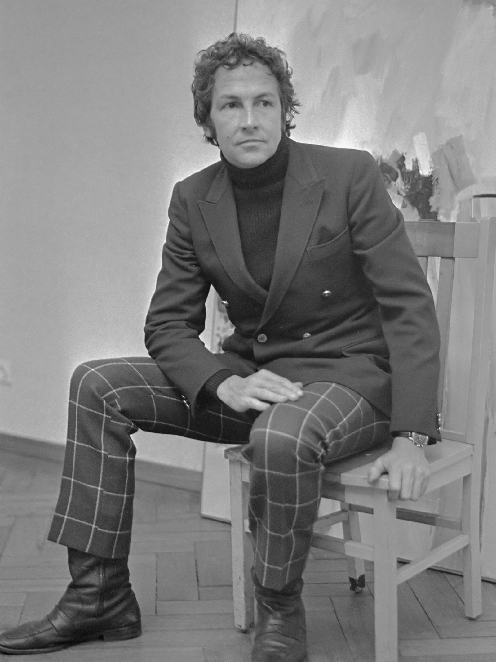 Black and white photo of Robert Rauschenberg in a dark jacket and checked trousers seated on a wooden chair