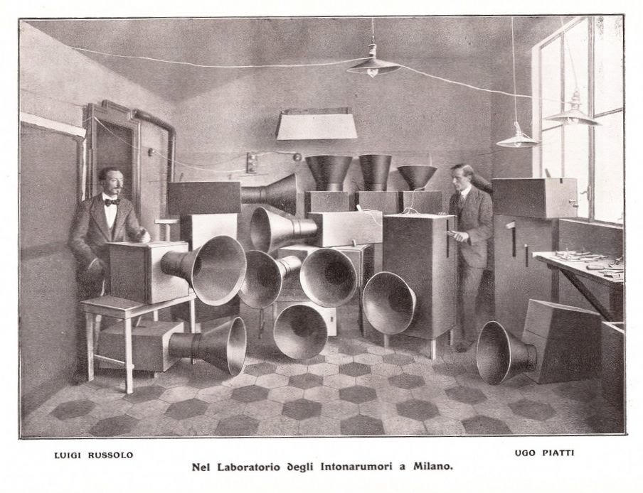 Black and white photograph of two men standing among stacked noise instruments with large horns in a workshop