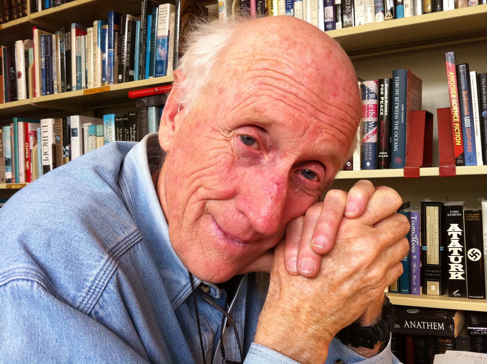 Stewart Brand in a blue shirt standing on the deck of a boat beside a red door, with the Sausalito waterfront behind