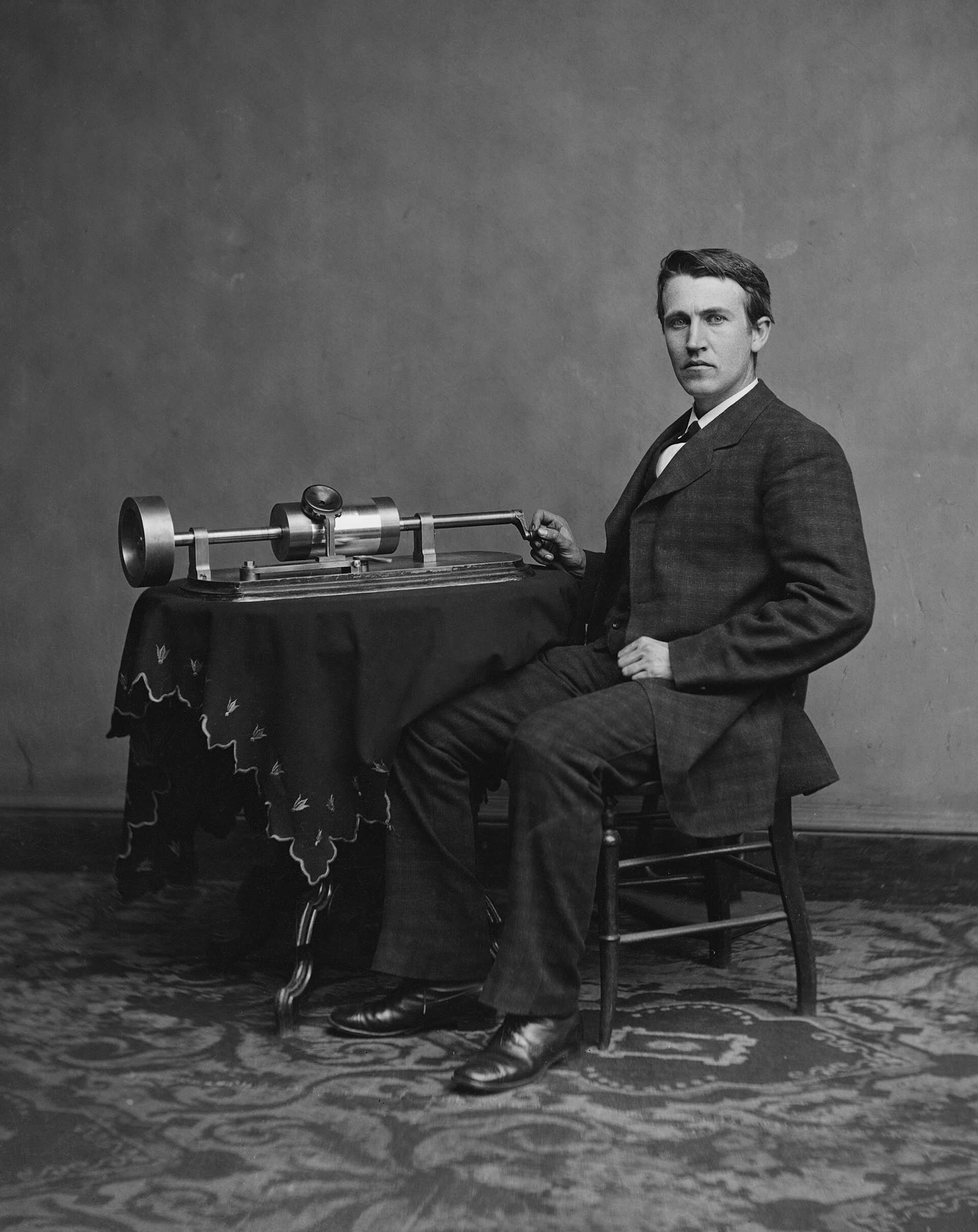 Studio photograph of Thomas Edison seated beside his early phonograph