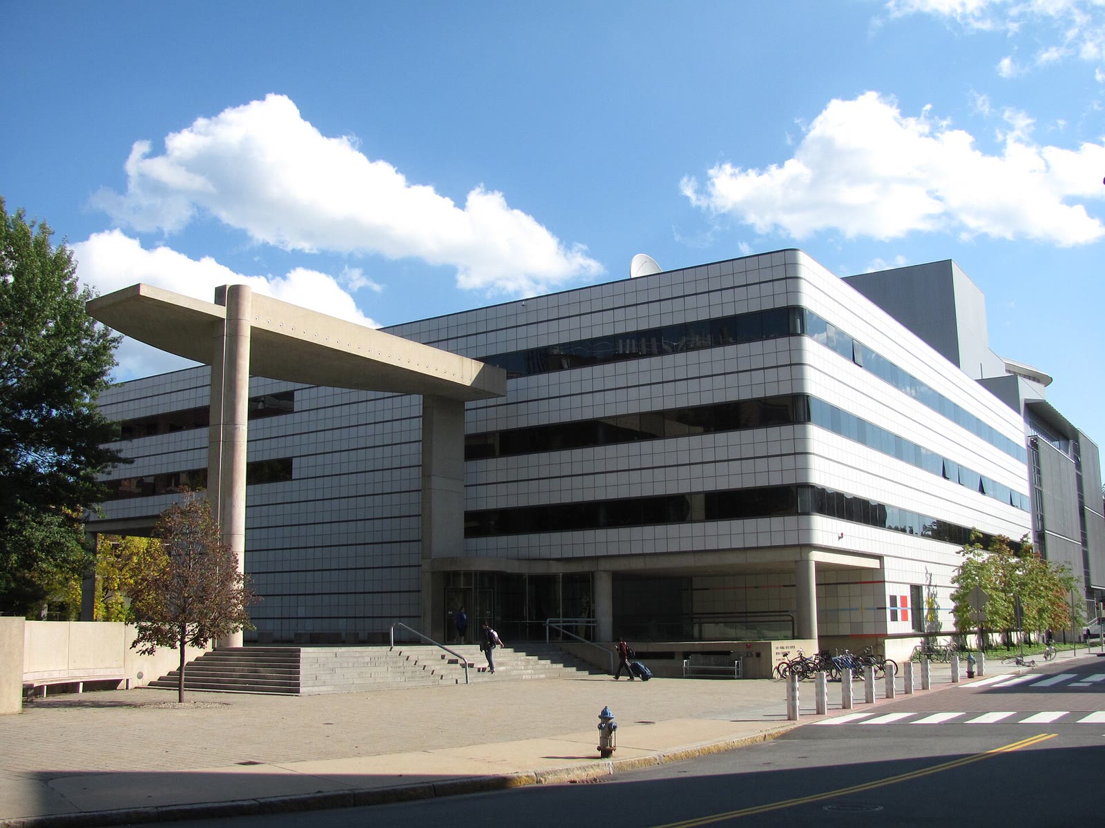 White tiled modernist Wiesner Building at MIT with a concrete portal frame in front, under a blue sky