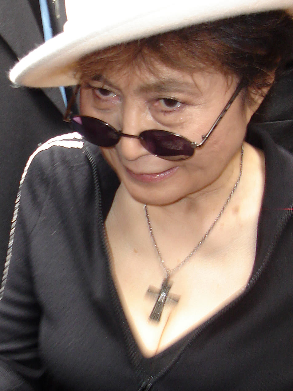 Yoko Ono in a white hat, dark glasses and a black top with a cross pendant, looking down